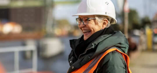 Woman in field services industry at job site, smiling, holding a tablet.